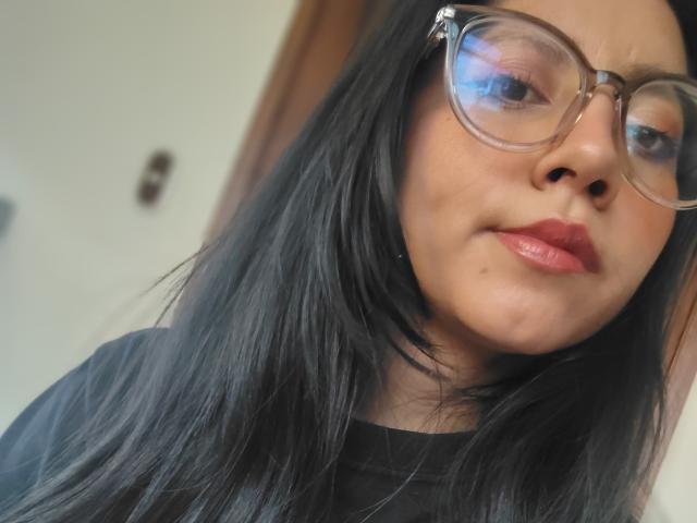 VioletXBloom heeft lang zwart haar, heldere glazen en glanzende rode lippen. Haar gezicht vertoont een lichte grijns met een kleine puist op haar linkerwang. Ze draagt een zwart shirt..