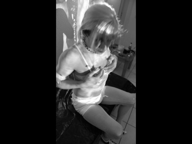Schwarz-weißes Foto von TVNutte-Hot mit blonden Haaren, die einen BH, eine Halskette und Fischernetzstrümpfe trägt und auf einem Stuhl sitzt, wobei sie ihre Brüste mit ihren Händen bedeckt..