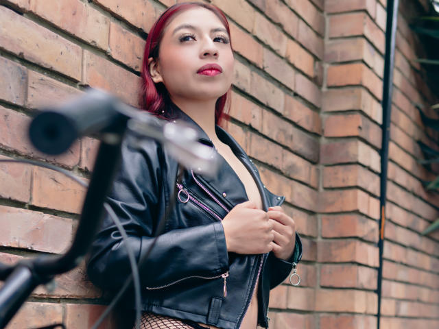 ZoeRedds mit roten Haaren, einer schwarzen Lederjacke und freiliegendem Brustbereich, hält ein Mikrofon vor einer Ziegelwand und zeigt einen selbstbewussten Ausdruck mit pinkfarbener Lippenstift..