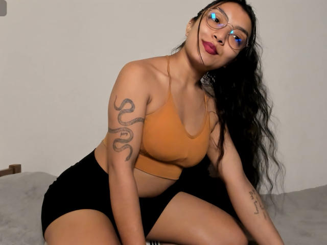 IbbyeMoreau with long black hair, glasses, orange sports bra, black shorts, snake tattoo on right arm, sitting on bed, looking at camera, pink lips, blue eye reflection.