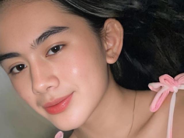 YurieFresh with smooth, fair skin, dark eyebrows, red lips, pink ribbon top, and a slight smile. Her black hair is pulled back, exposing her delicate features.
