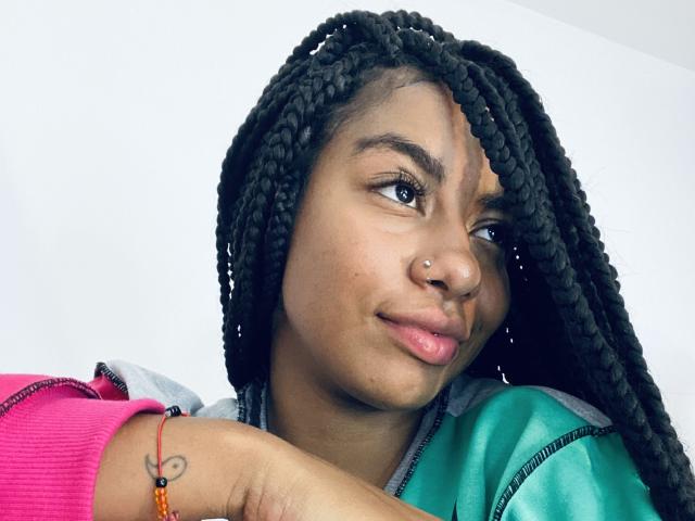 AhteneaSexy, com tranças escuras, lábios levemente entreabertos e um piercing no nariz, vestindo uma jaqueta verde e rosa, mostrando uma tatuagem de coração no braço. Ela está com uma expressão adorável..