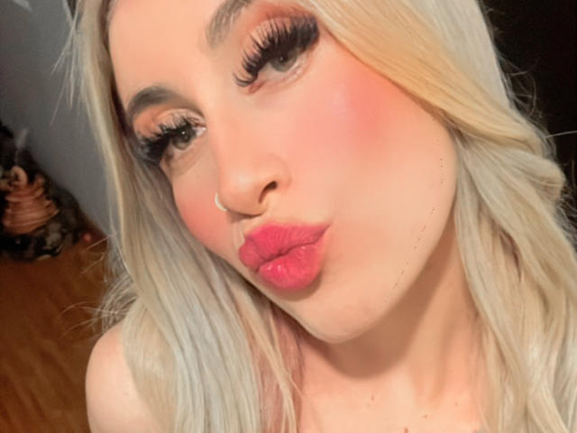 SammyViolet, met haar platinablonde haar, roze blush en glanzende roze lippen, trekt haar mond op. Ze heeft dikke, lange wimpers, een kleine neusring en een licht omhoog gekeerd, schattig gezicht..