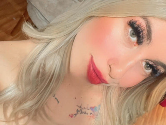 SammyViolet, com cabelo loiro, blush rosa, batom vermelho e um piercing no nariz, fazendo beicinho com os seios levemente visíveis, tatuagens no peito e cílios longos em um selfie..