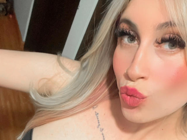 SammyViolet with pouty pink lips, blonde hair, and long lashes, making a kissy face, showing a tattoo on her upper chest, with her arm raised.