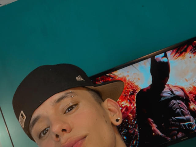HardinBat, wearing a black cap, has a small facial piercing, black ear gauges, and a slight smile, with a Batman movie poster in the background.