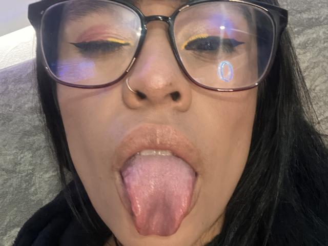 StellaBeckker's face, tongue out, wearing black-framed glasses with yellow eyeliner, reflection on glasses, nose ring, black hair, gray background. Boobs not visible. Ass not visible. Pussy not visible.