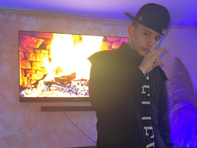 HardinBat with short brown hair, black cap, black shirt with white straps, holding finger to chin, standing in front of a wall-mounted TV showing a colorful fire image.