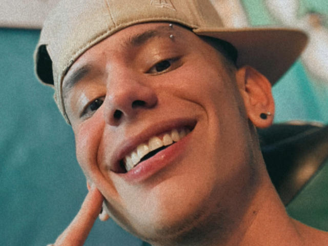 HardinBat's smiling face with a small nose piercing, wearing a beige baseball cap, and a black earring, showing off a fresh set of white teeth and a cute butt cheek.