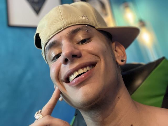 HardinBat's smiling face, wearing a beige cap, has a nose piercing and a ring on his finger, with a blue background and blurred lights in the background.