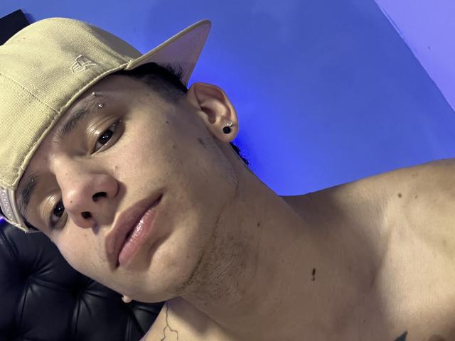 HardinBat with a light beige baseball cap, shirtless, showing a nose piercing, black ear hole, and a slight smile, blue-lit background, short dark hair, and a tattoo on the neck.