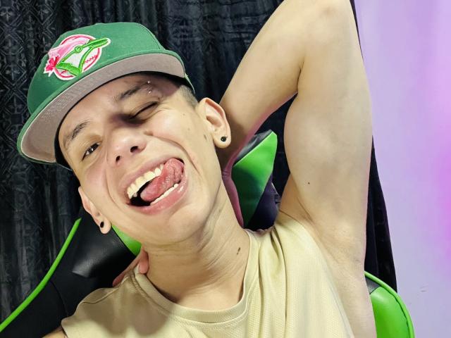 HardinBat with a green hat, winking, tongue out, smiling, showing armpit, small earrings, and a tan shirt, sitting in a green and black chair.