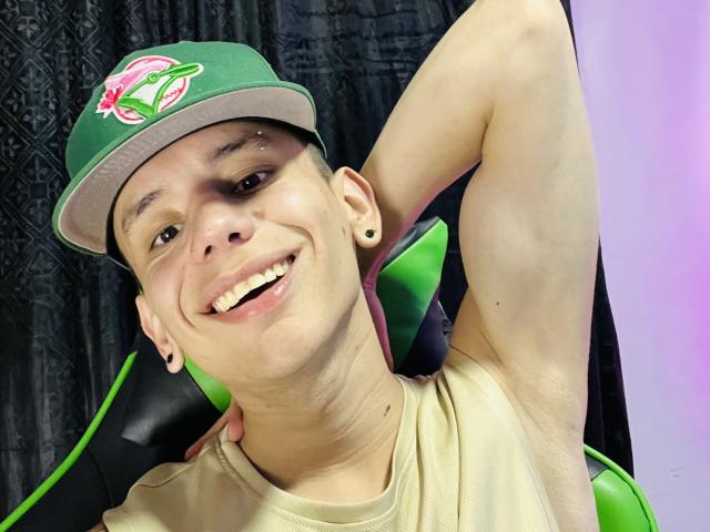 HardinBat with short brown hair, green cap, and ear gauges, smiling with arm behind head, wearing beige shirt, sitting in green-black chair. Visible armpit, black earbuds.