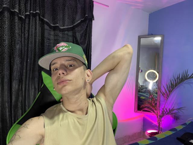 HardinBat, wearing a green cap with a pink flower, beige sleeveless shirt, arms raised, tattoo on left shoulder, sitting in a green-black chair, mirror with ring light behind.
