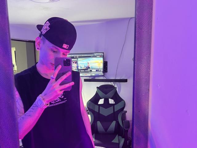 HardinBat with tattoos, wearing a black cap and sleeveless shirt, taking a mirror selfie with purple lighting. Gaming chair and TV in the background.