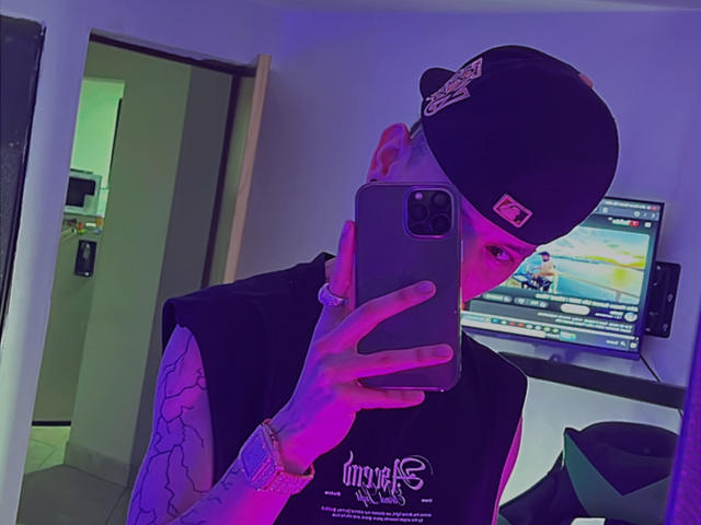 HardinBat with tattoos, wearing a black cap and sleeveless shirt, holding a phone in front of his face, purple and blue lighting, reflection in mirror.