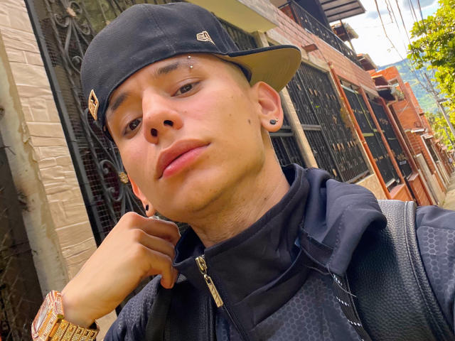 HardinBat with a black cap, nose piercing, and black hoodie, holding his chin, wearing a gold watch, and sporting ear studs, looking into the camera with a slight smirk.