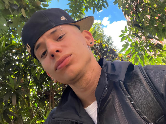 HardinBat with a black cap, nose piercing, and earring, wearing a black jacket, looking down with a slight smirk, surrounded by green leaves and a blue sky.