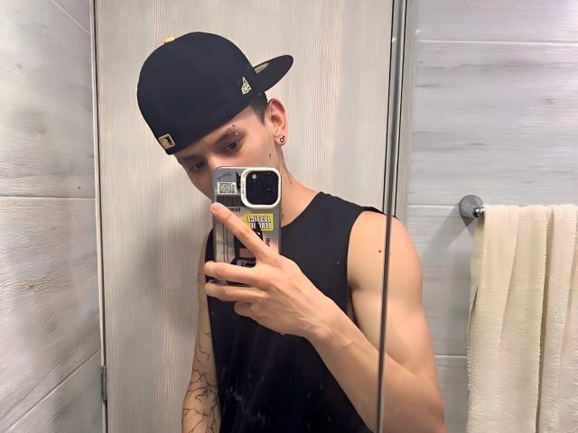 HardinBat with a black cap, sleeveless black shirt, tattooed arm, and ear piercings, taking a mirror selfie in a bathroom, holding a phone with a yellow sticker.