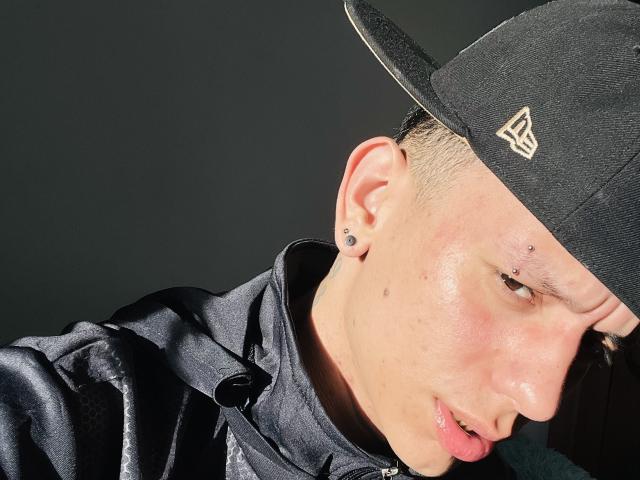 HardinBat with a black cap, ear studs, and a nose ring, wearing a shiny black jacket, sunlight highlighting his face and slightly blushing skin.