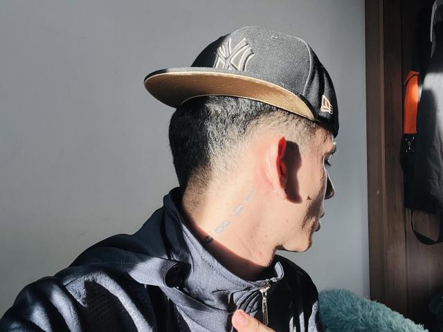 HardinBat's side profile, wearing a black NY cap with a gold brim, black zip-up jacket, and visible neck tattoo, with sunlight highlighting his short dark hair and mustache.