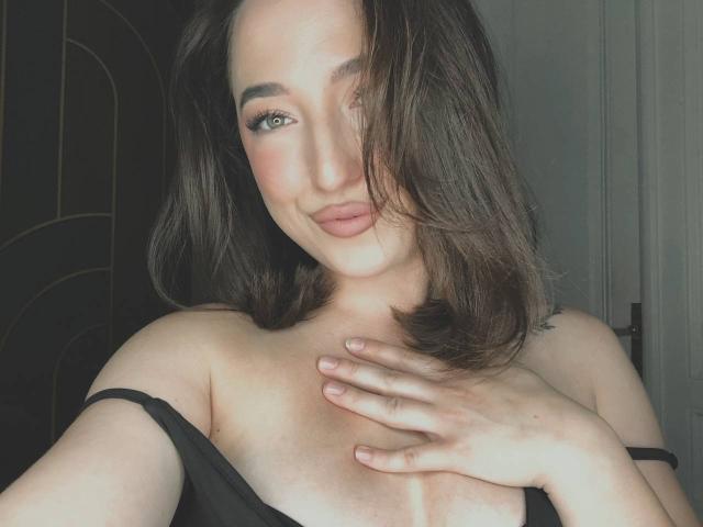 MonAmourR met bruin haar tot op de schouders, groene ogen en roze lippenstift, die zachtjes glimlacht, met haar hand op haar borst, een zwarte band die van haar afglijdt, waardoor een beetje van haar borsten zichtbaar zijn..