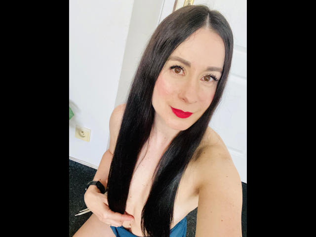 HeisseVicki-hot with long black hair, red lipstick, and blue dress, holding her tits, smiling, wearing a black wristband, in a bright room.