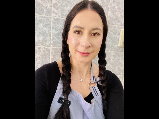 HeisseVicki-hot has braided black hair, a light skin tone, and is wearing a black cardigan with a gray apron, smiling with pink lipstick. She's a cute bitch.