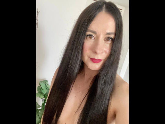 heisseVicki-hot with long black hair, red lipstick, and medium tits, smiling at the camera, topless, with a green plant in the background.