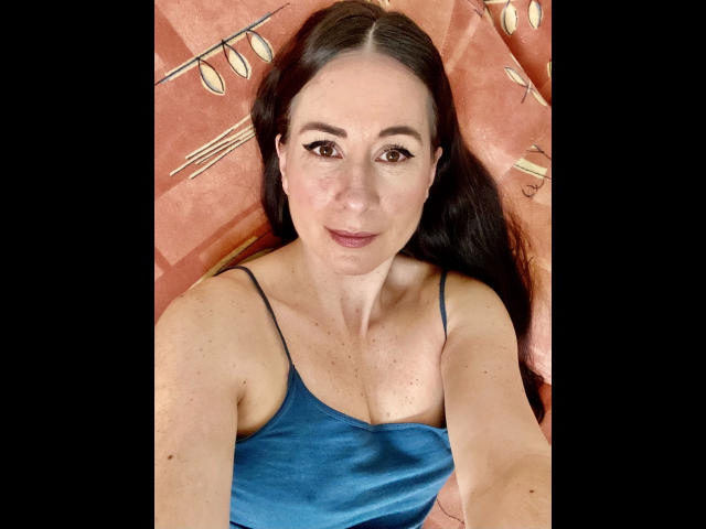 HeisseVicki-hot with long dark hair, wearing a blue satin tank top, showing her tits and a slight smile, lying on a pinkish patterned surface.