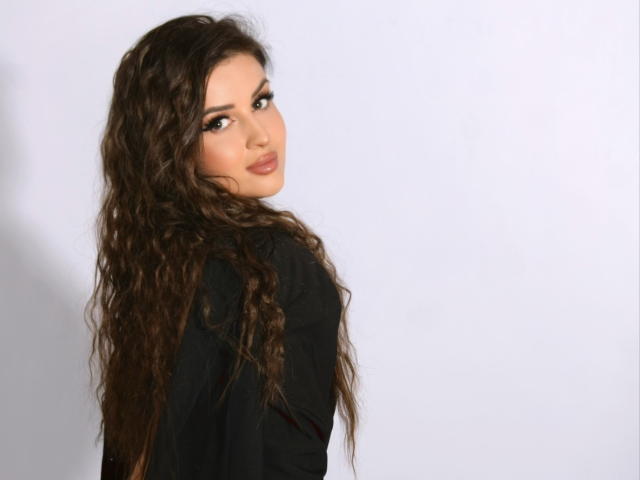 JessieAmore, con el pelo largo y ondulado de color castaño, mirando por encima del hombro, lleva una blusa negra, tiene labios carnosos y una ligera sonrisa, sus ojos son marrones y el maquillaje es discreto..