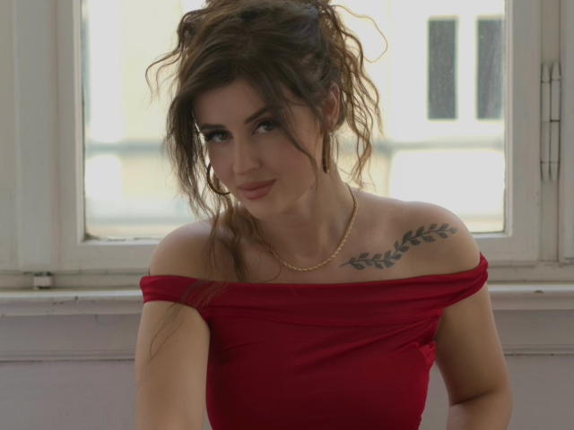 JessieAmore, avec ses cheveux bruns ondulés, sa robe rouge à une épaule, son collier en or et son tatouage en forme de feuille sur l'épaule droite, a l'air adorable avec un léger sourire, et se tient devant une fenêtre..