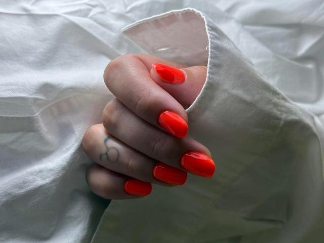 SarahFontain's hand with bright red, glossy nails, holding her white shirt sleeve. She's got a small tattoo on her index finger. Soft, natural light.
