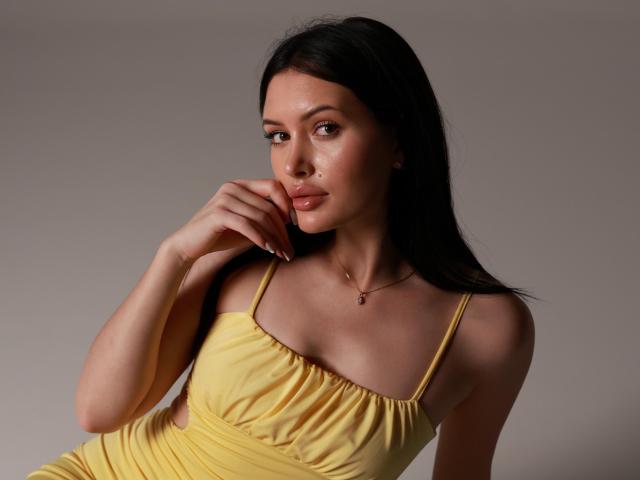 MilanaMusee with long black hair, yellow dress, and a hand near her pouty lips, wearing a necklace, has small tits and a cute face, looking seductive.