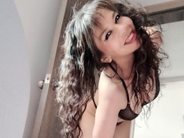 SamaraBellet with curly black hair, smiling, wearing black lingerie, bending forward, showing small, perky tits, and a cute face, in a selfie with a playful, sexy expression.