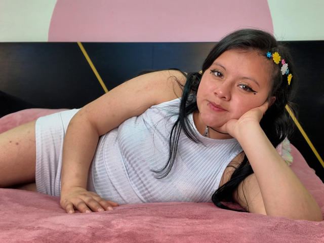 KatteReed, a chubby woman with long black hair, wearing a white ribbed tank top, lying on a pink bed, head resting on hand, small flowers in hair, smiling.