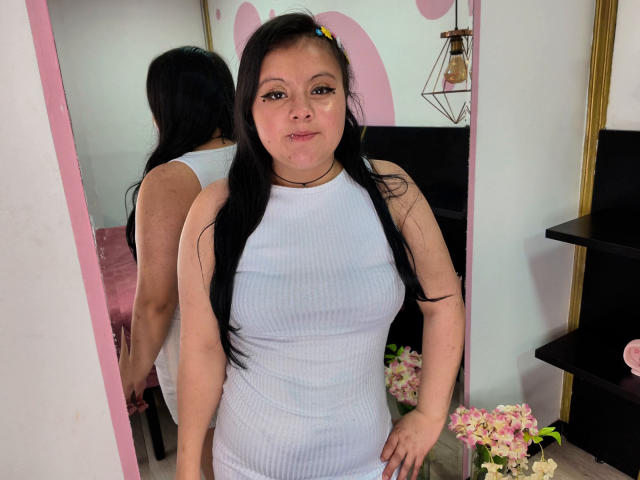 KatteReed with long black hair, wearing a white ribbed dress, standing in front of a mirror, showing her tits and ass in the reflection. Fluffy pink and white flowers.