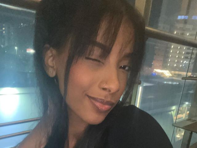 StormiBlaze has a cute, winking smile with shiny pink lips, a small nose ring, and dark, messy hair. She's wearing a black top, looking sexy and adorable.