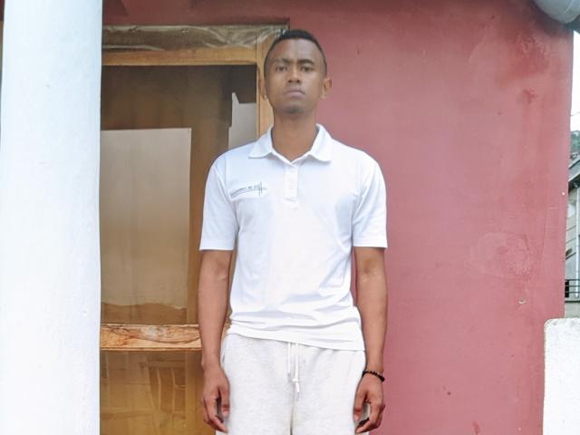 SugarBoyd stands with a serious face, wearing a white polo shirt and white pants, hands in pockets, brown bracelet on right wrist, against a red and white wall.