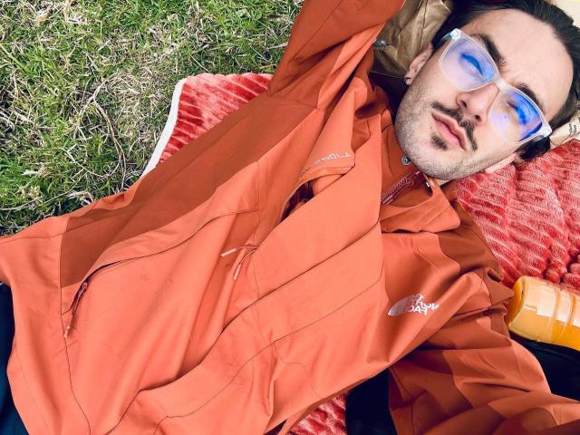 JakeMagnus, with orange jacket and reflective sunglasses, lying on red blanket, arms above head, looking relaxed, holding orange bottle, grassy ground, short dark hair, slight beard, casual vibe.