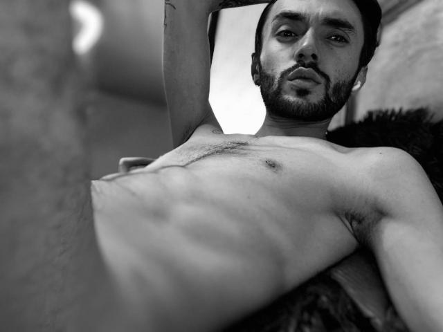 JakeMagnus, shirtless, lying down, arms raised, looking at camera, displaying a slim, hairy chest with visible abs and small nipples, black and white, semi-erect penis in foreground.