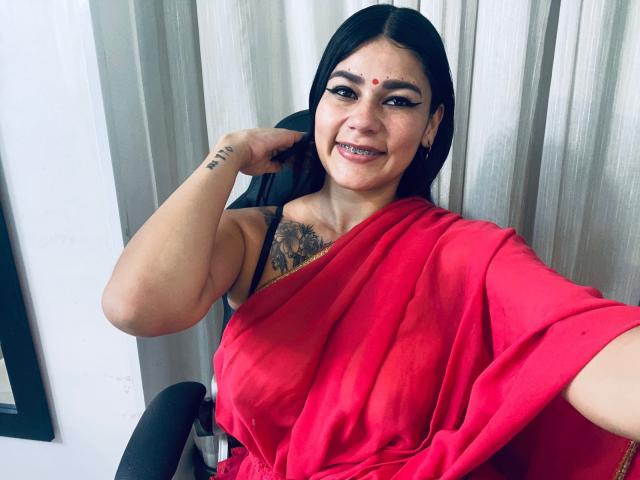 KeraniMarala met zwart haar, een rode sari en een tatoeage op de borst, lachend, hand op het hoofd, zittend in een stoel, met de borsten zichtbaar, een kleine rode stip op het voorhoofd, ontspannen, selfie..