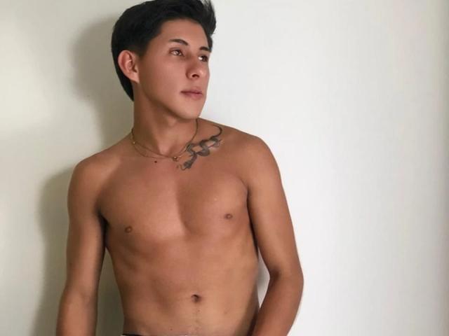 SoffiaVegga, shirtless with a lean, muscular build, small nipples, and a tattoo on the right chest. Short dark hair, looking to the side, wearing a thin gold necklace.