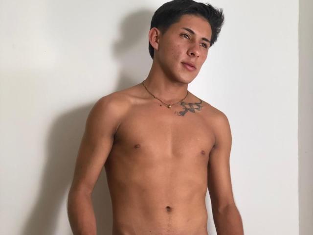 SoffiaVegga is a shirtless, tan-skinned guy with short black hair, a small chest tattoo, and a necklace, standing against a white wall. His cock is not visible.