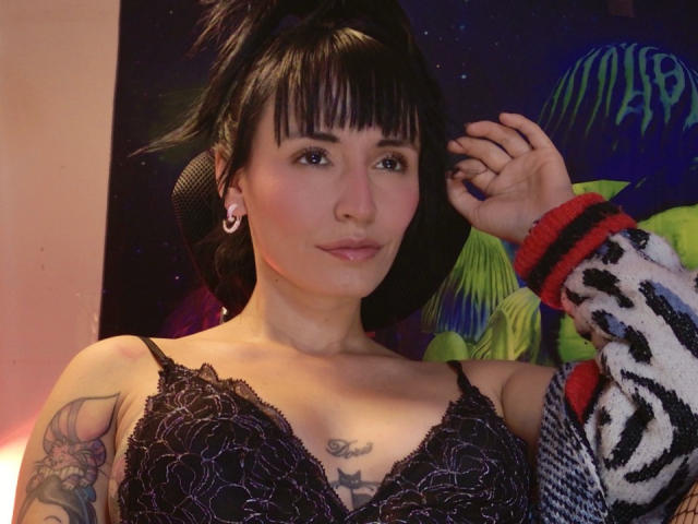 SaraleePoison met een zwarte kanten BH, kleine borsten, tatoeages op de rechterarm en borst, zwart haar in een hoge paardenstaart, draagt armwarmers met een zebrapatroon, oorbellen en subtiele make-up..