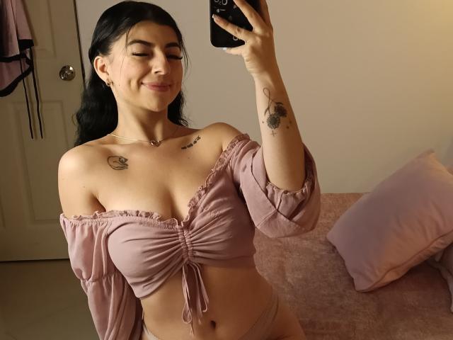 AnntonellaWatson with black hair, smiling, holding a phone, wearing a pink off-shoulder top, showing small tits, tattoos on chest and arm, pink panties, in a bedroom.