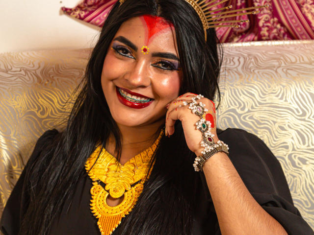 LakshmIrani with black hair, red forehead dot, gold headpiece, yellow necklace, black dress, red lipstick, smiling, showing teeth, hand with jewelry, sitting, dark skin, attractive.