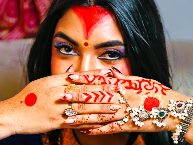 LakshmIrani's hands with red henna and rings cover her mouth, showing off her painted nails and a red dot on her forehead, with dark hair and purple eye makeup.