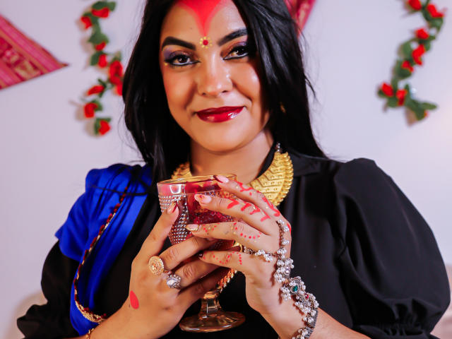 LakshmIrani with dark hair, red makeup, and gold necklace holds a red glass cup. She's wearing a blue and black outfit, smiling, with red henna on her hands.