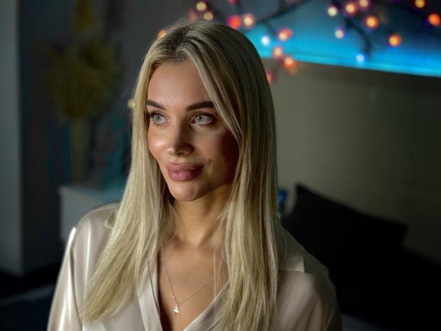 SabrinaSharm with blonde hair, blue eyes, and pink lipstick, wearing a shiny white blouse, and a small necklace, smiling gently. Boobless, no visible ass or pussy.